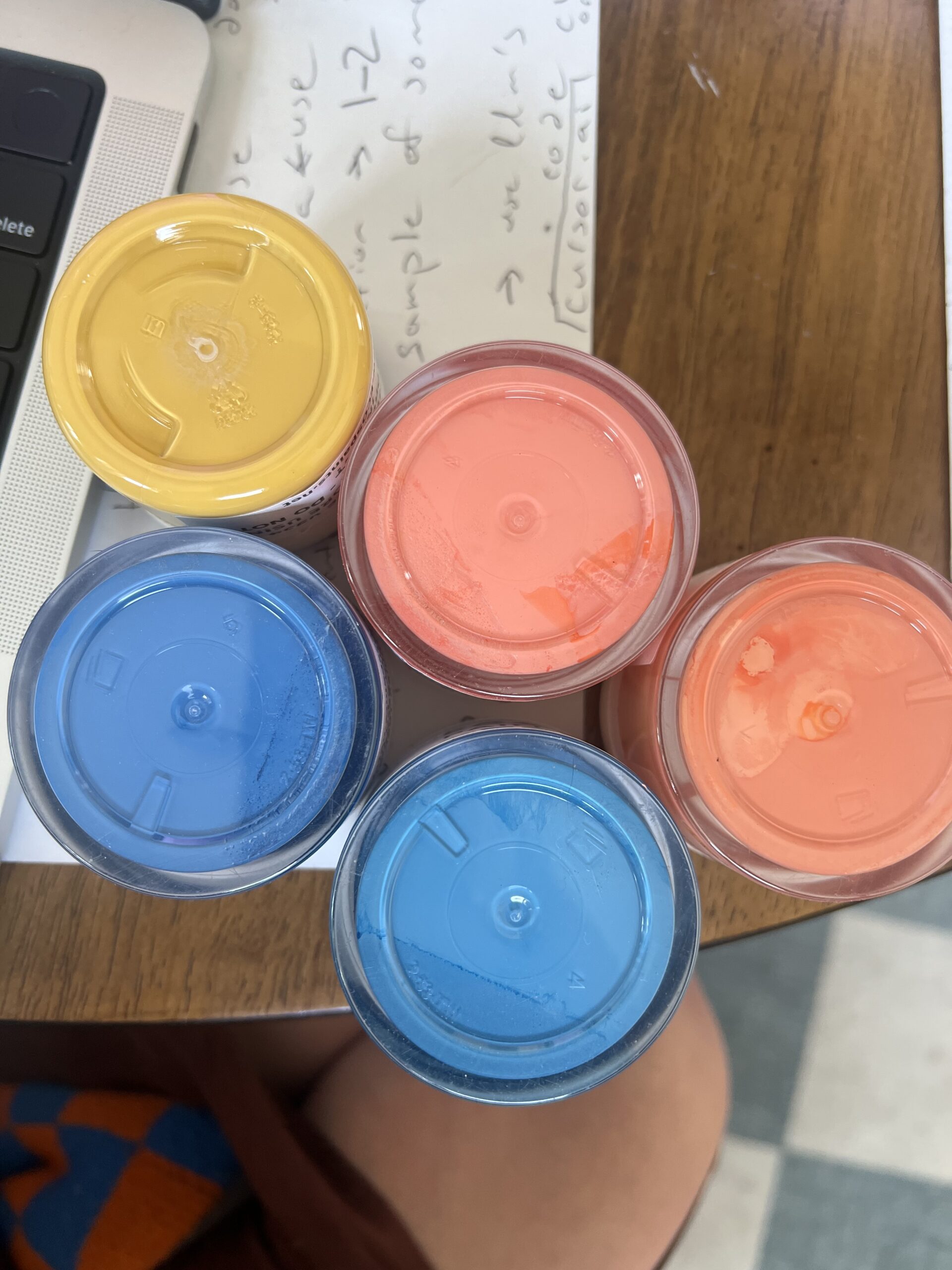 Five plastic containers with yellow, blue, and orange lids are arranged in a cluster on a wooden surface next to a laptop and a handwritten note.