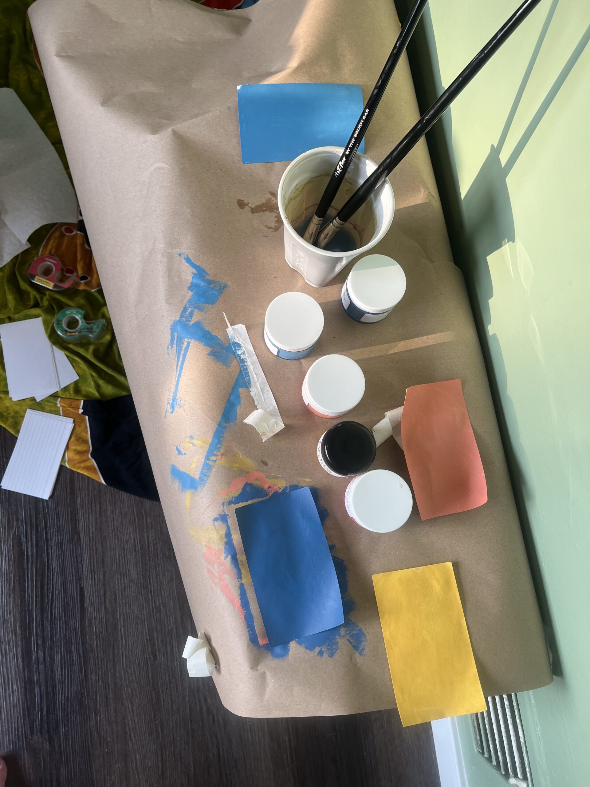 Art supplies including paint jars, brushes in a cup, and colored paper sheets on a paper-covered surface, with paint marks visible.