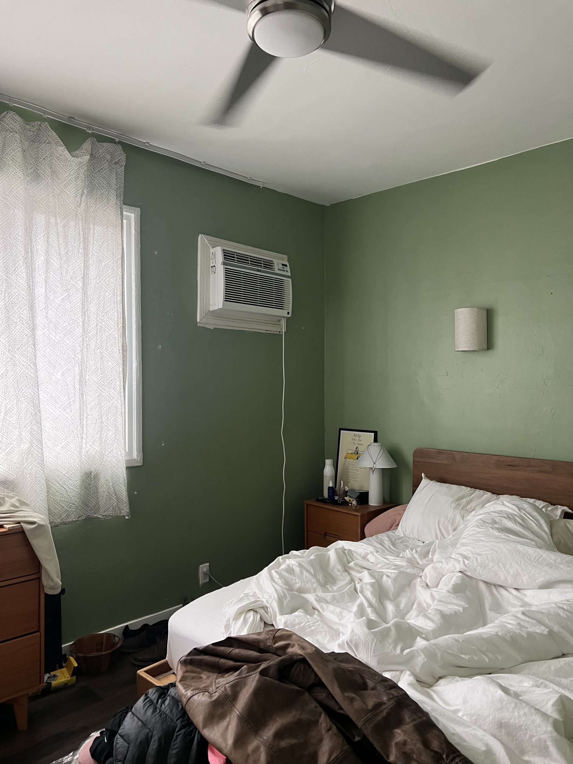 A bedroom with green walls, an unmade bed, wooden furniture, an air conditioner in the window, and light streaming through sheer curtains.