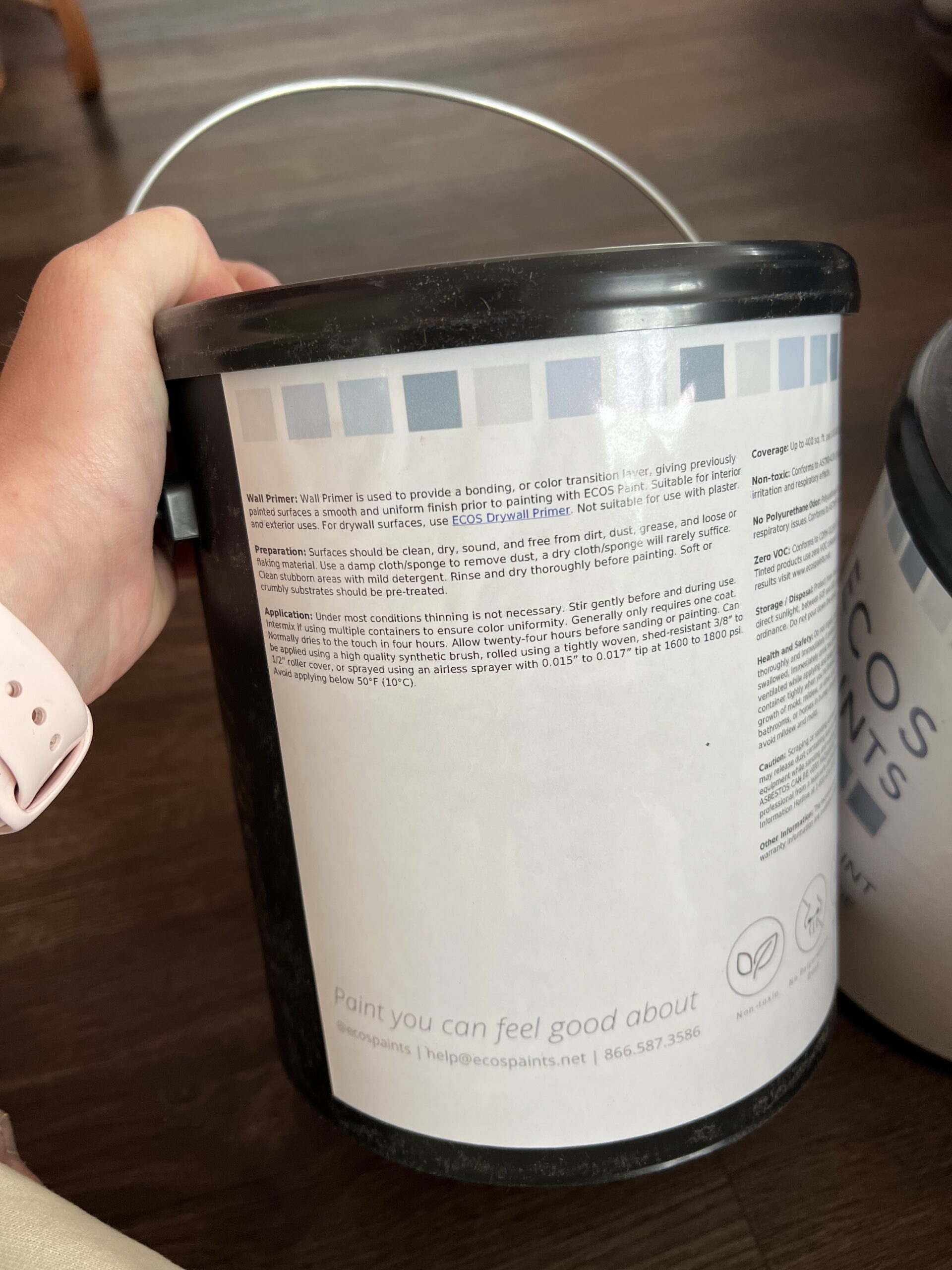 A hand holds a black and white paint can labeled "Ecos Paints" with detailed product usage instructions printed on the back.