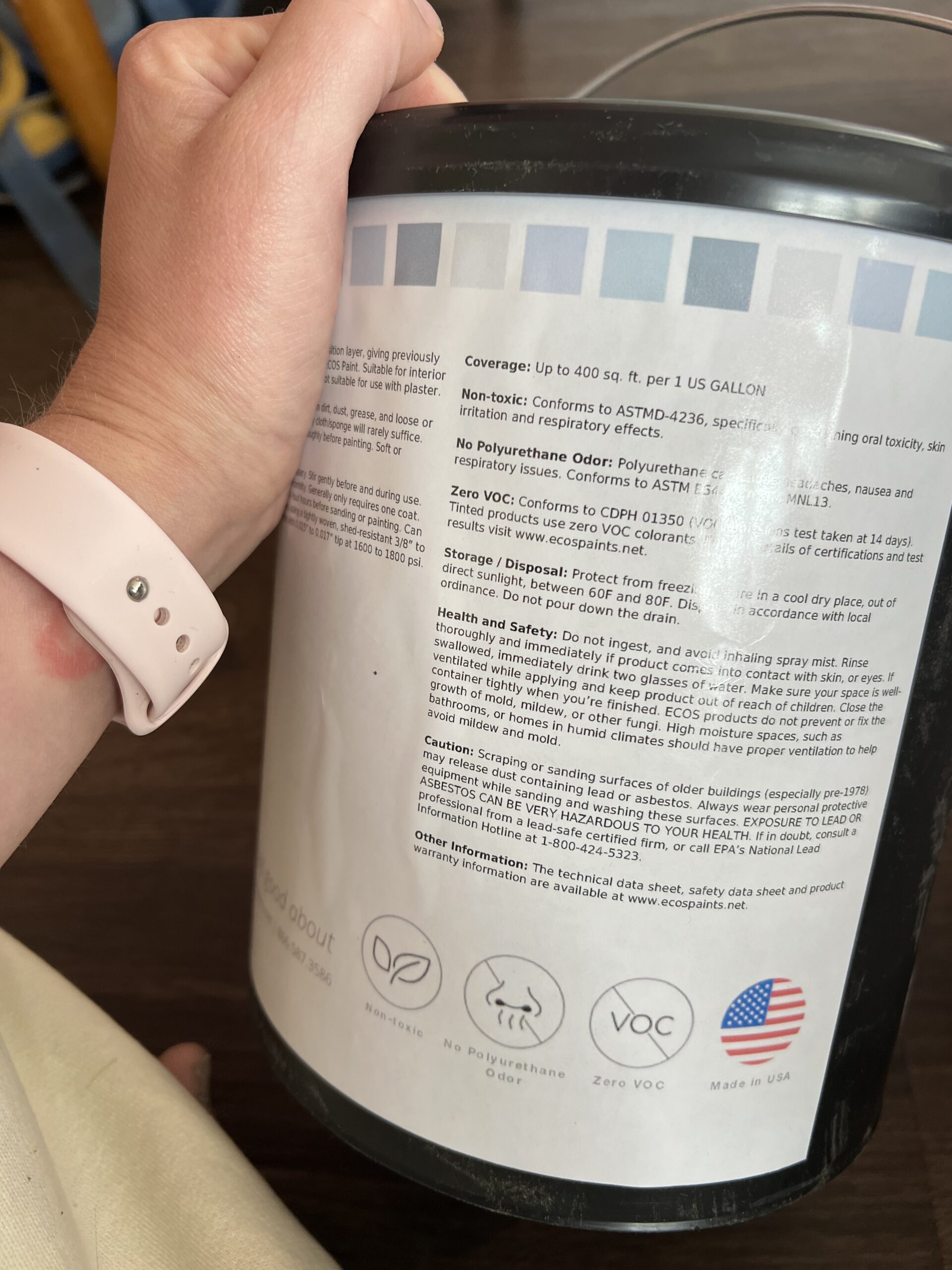 A hand holding a can of paint, showing the label with coverage details, safety instructions, and eco-friendly certification icons.