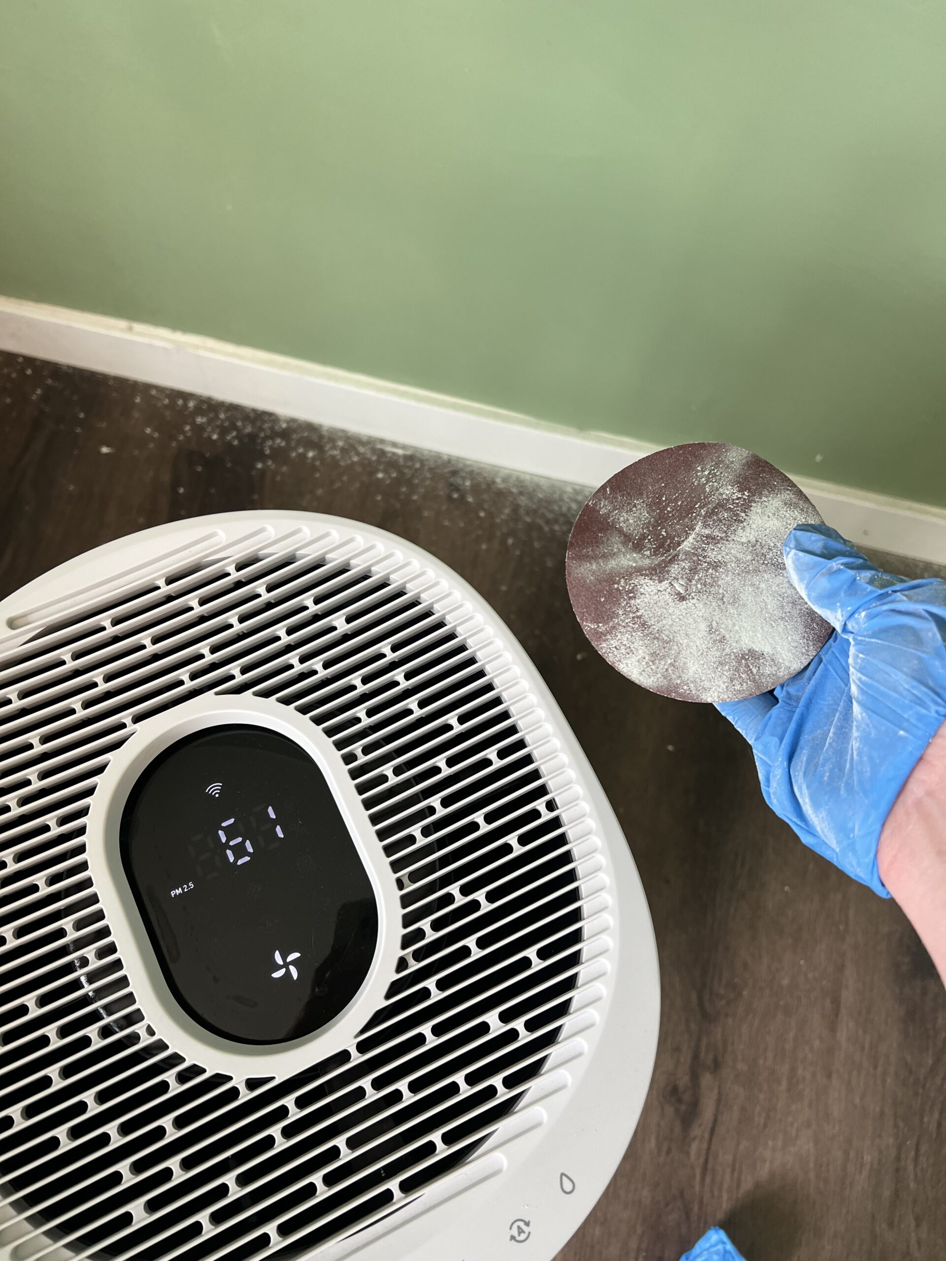 A gloved hand holds a dust-covered sanding disc near an air purifier on a wood floor with white dust scattered nearby.