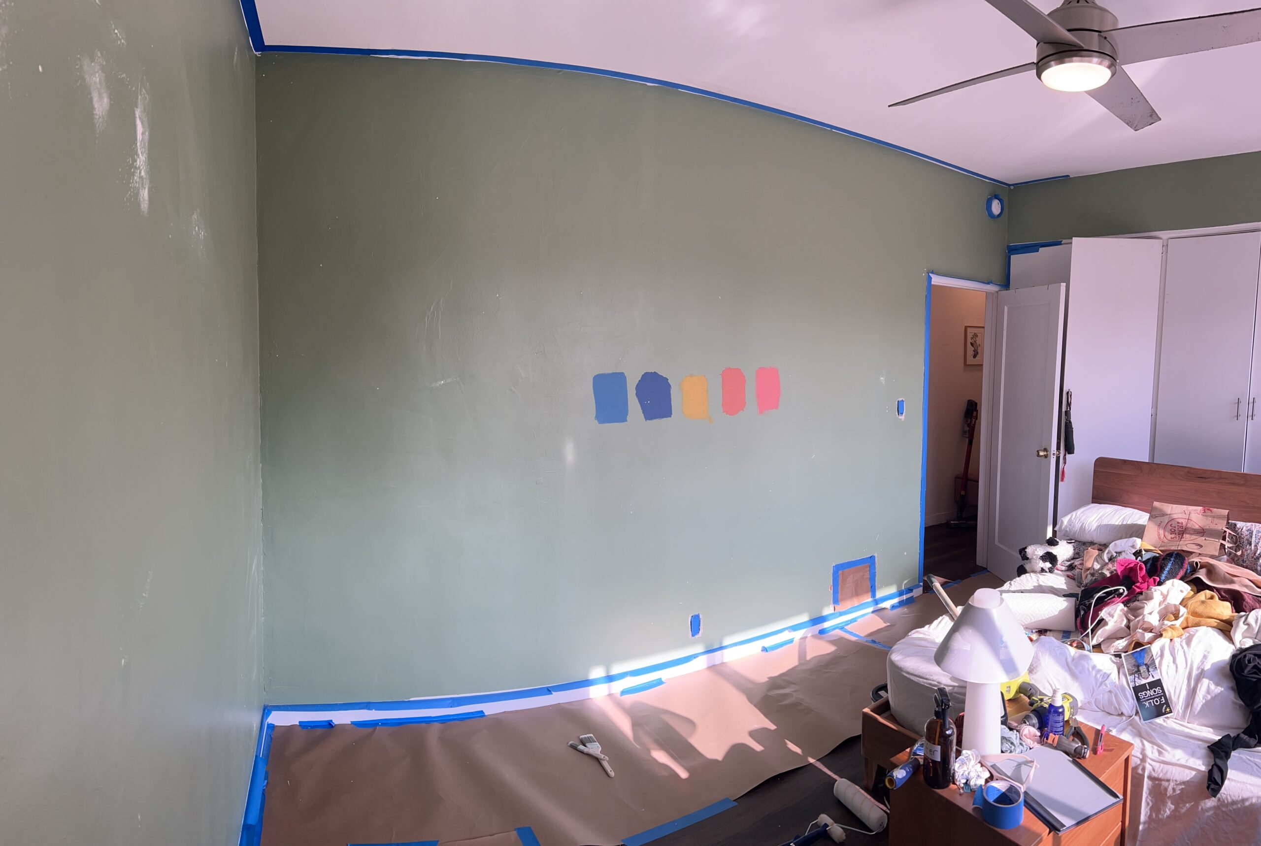 A green-walled room with blue painter’s tape along the edges, paint samples in blue, beige, pink, red, and yellow on the wall, and furniture covered for painting.