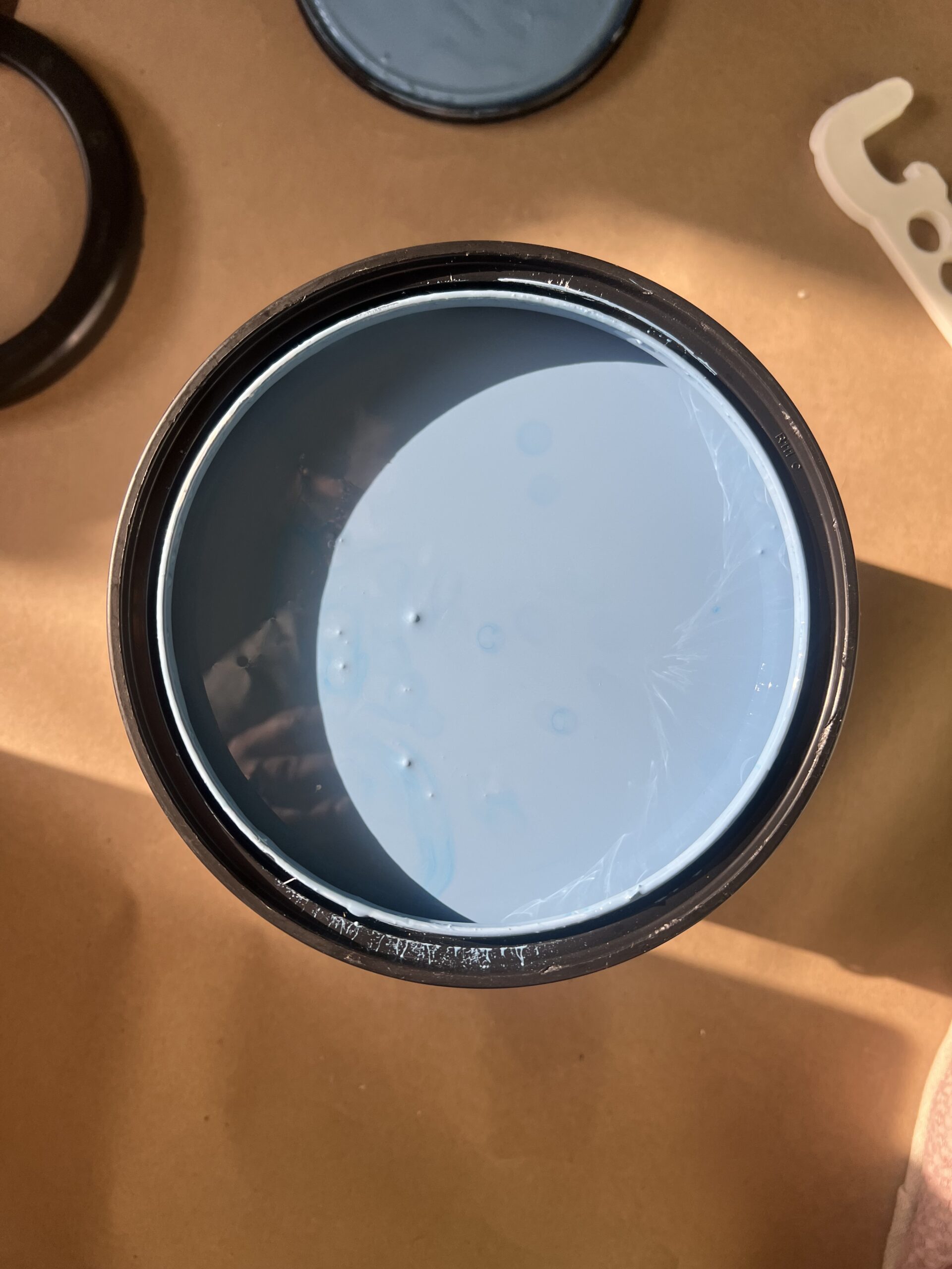 An open can of light blue paint sits on a brown surface, with the lid and a paint can opener nearby.