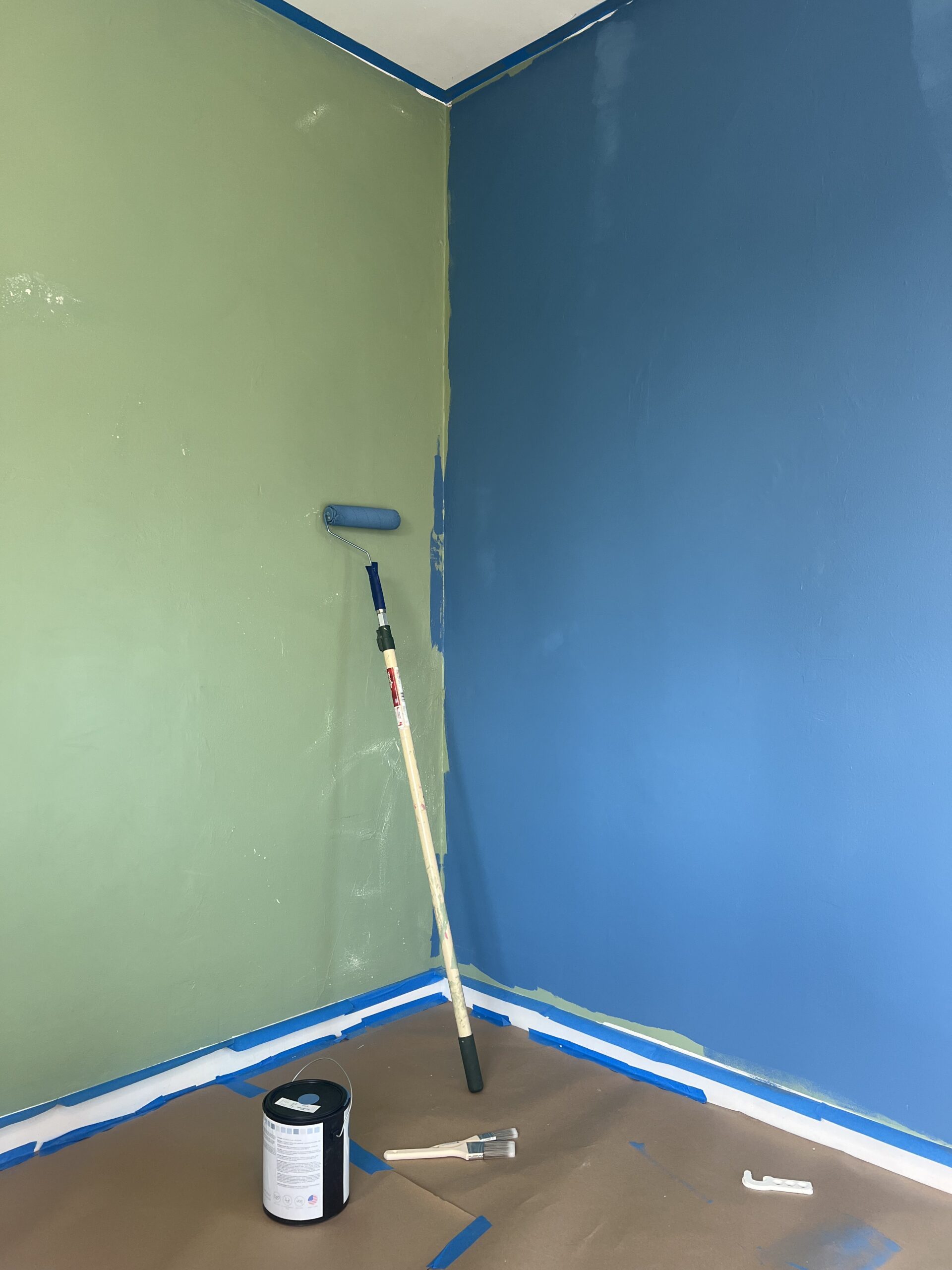 A room corner with one wall painted green and the other blue; a roller with blue paint leans against the green wall, and painting supplies are on the floor.