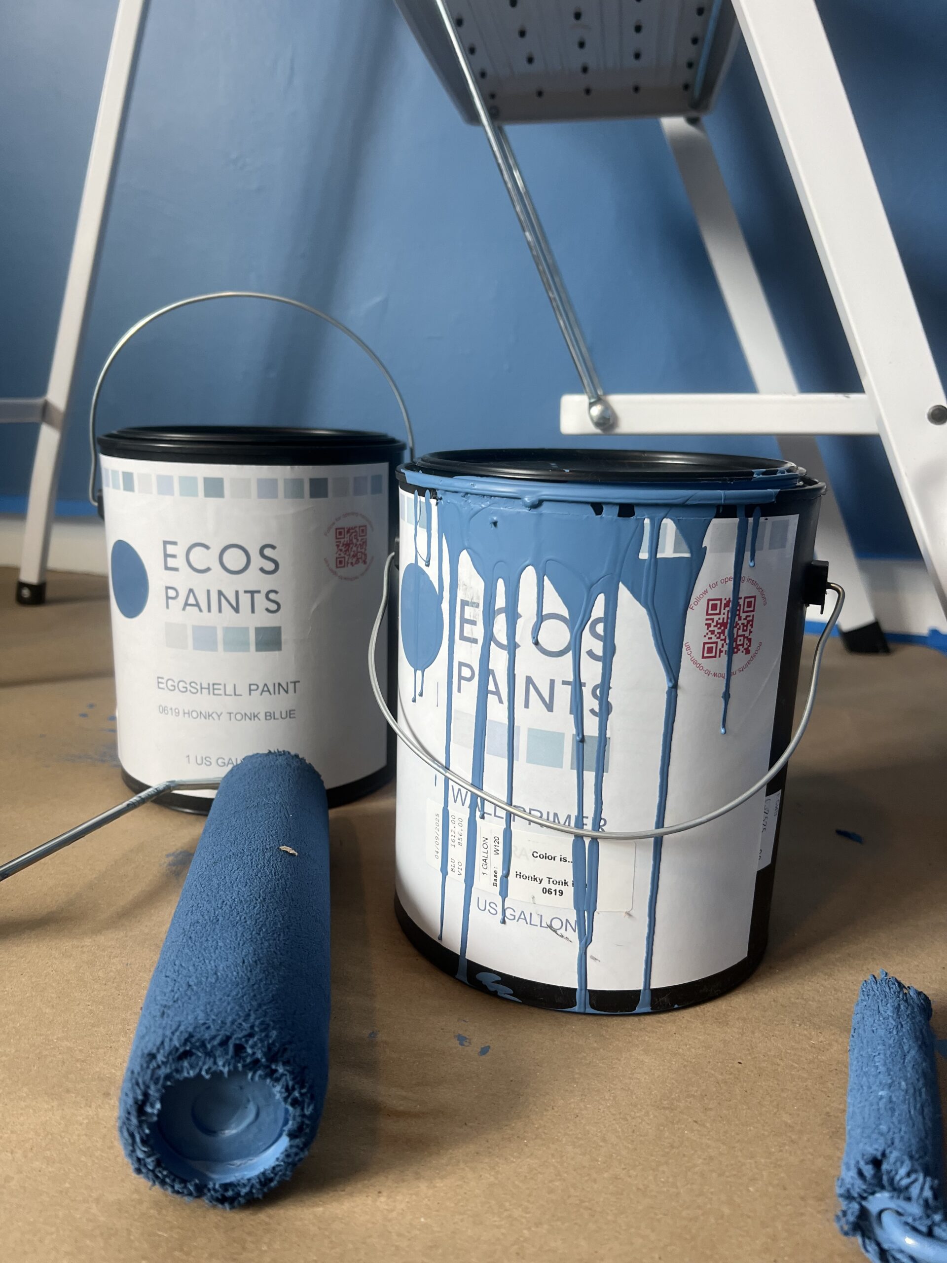 Two cans of ECOS blue eggshell paint, one with paint dripping down the side, are on a covered floor next to two paint rollers and a ladder, with a blue wall in the background.