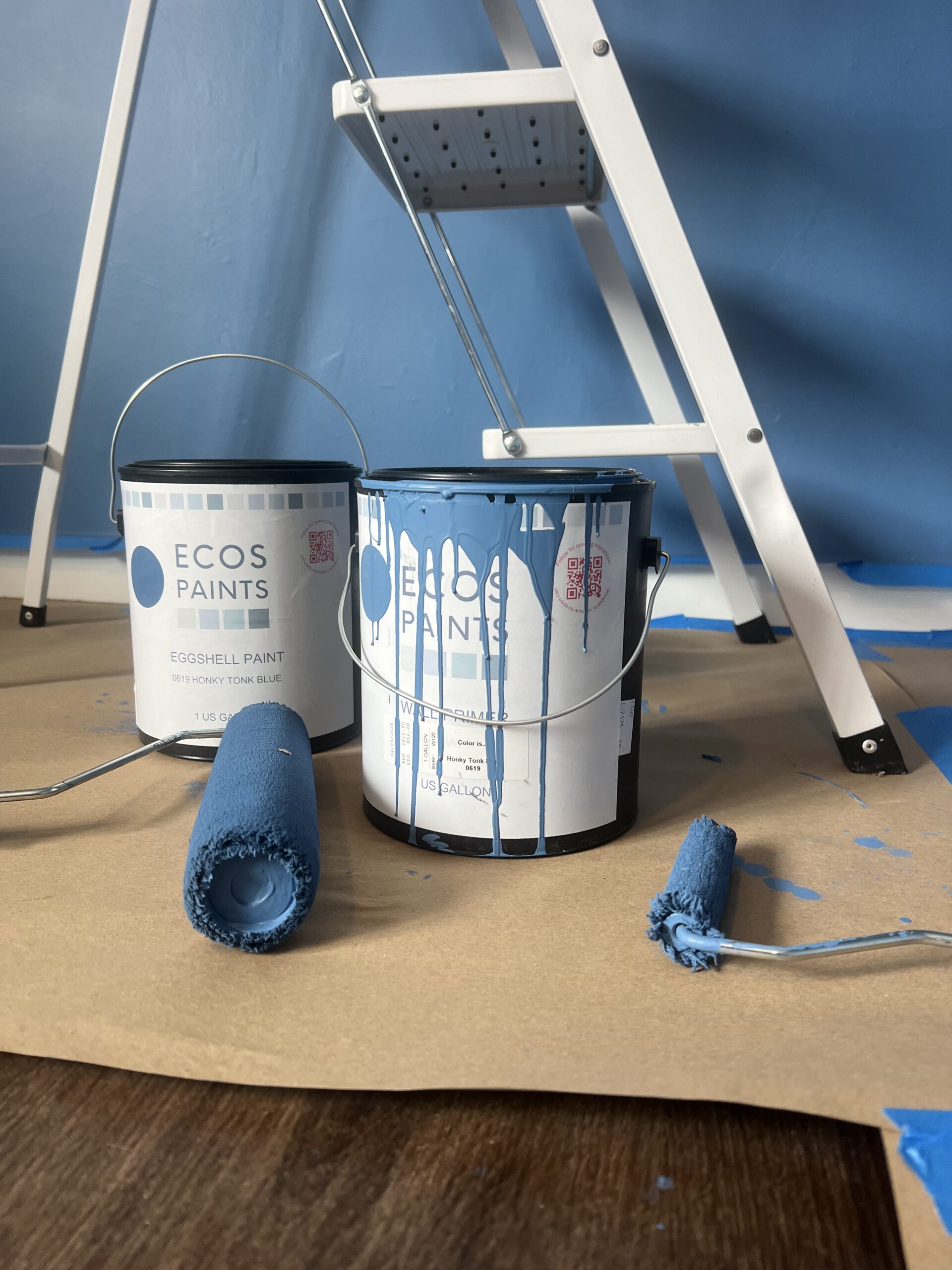 Two cans of blue ECOS paint, a paint roller, and a step ladder stand on a paper-covered floor with blue paint on the roller and splattered on the paint cans.