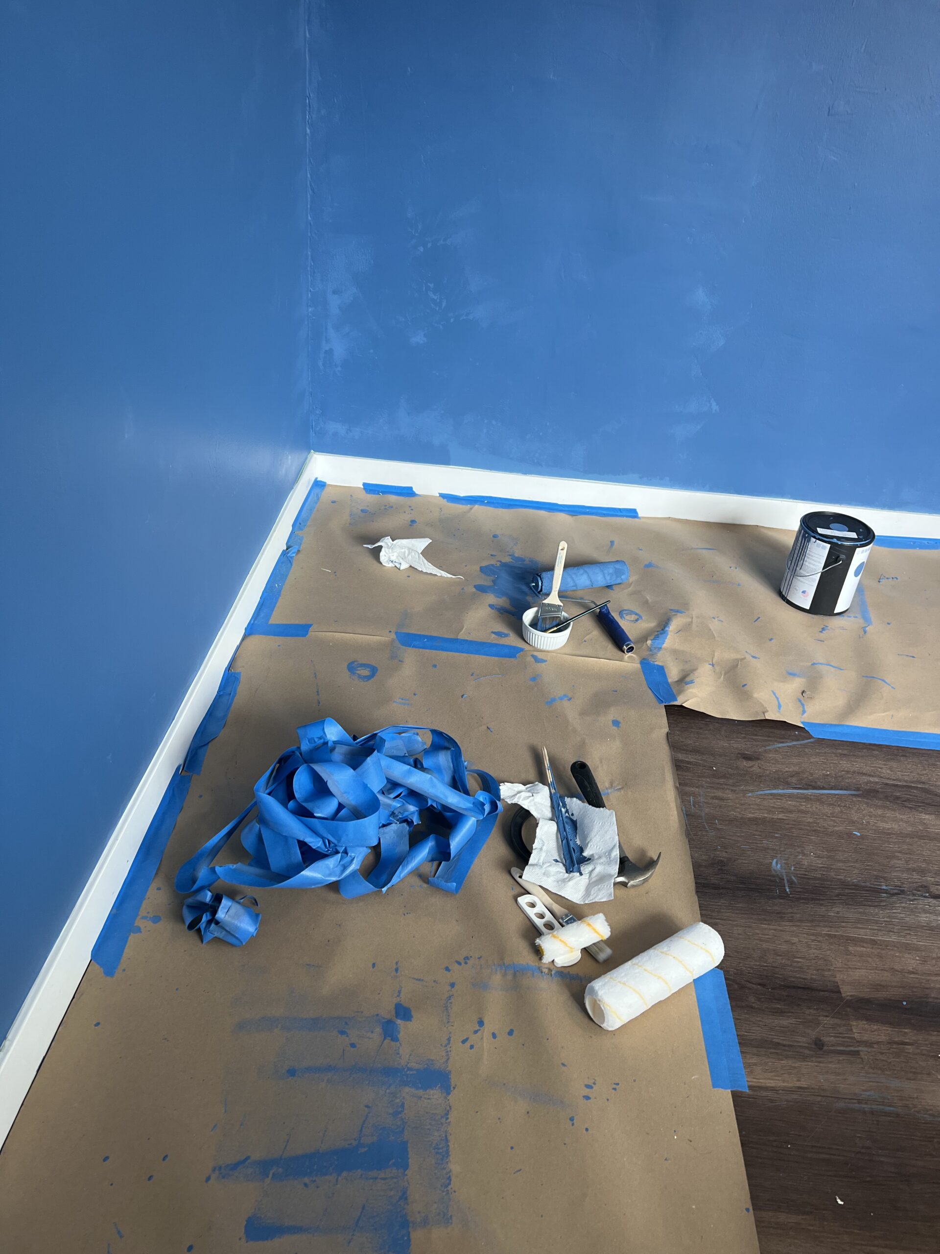 A room corner with blue-painted walls, painting supplies, tape, and paper covering the floor. A paint roller, brush, and paint can are visible on the floor.
