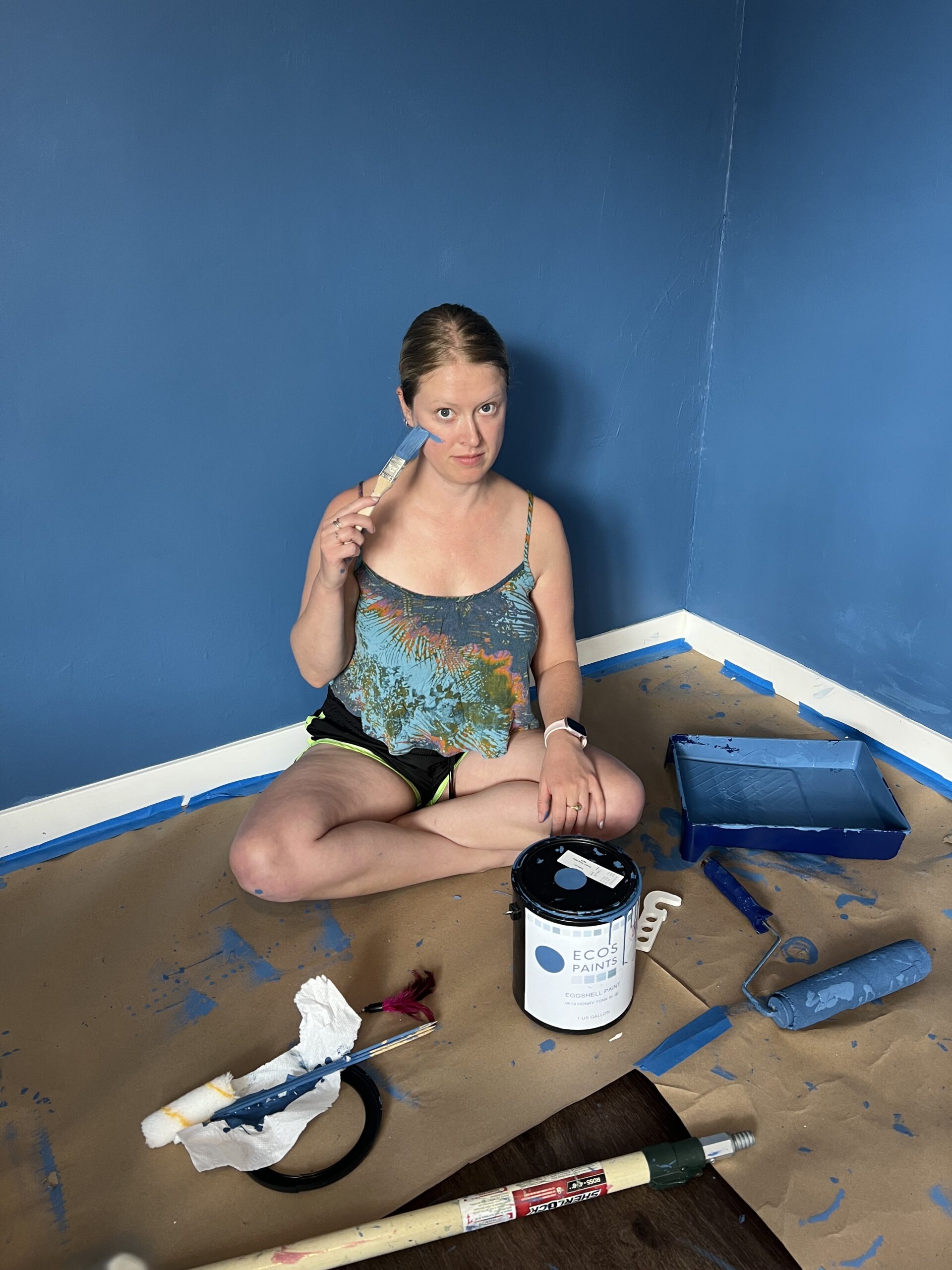 A woman sits on the floor of a room with blue walls, holding a paintbrush next to an open can of blue paint and painting supplies on a paper-covered floor.