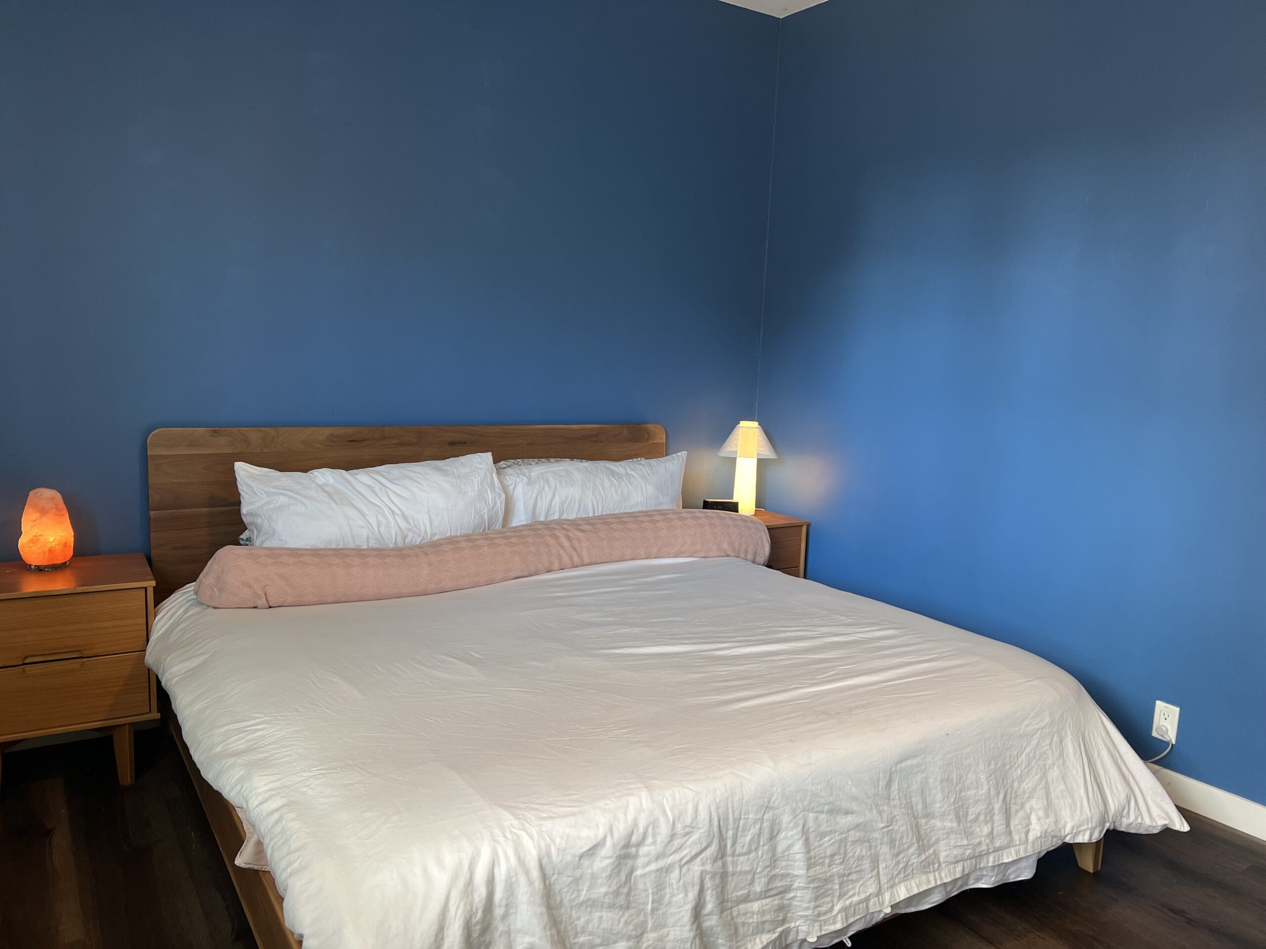A neatly made bed with white bedding and a pink pillow sits against a blue wall. Two wooden nightstands hold a salt lamp and a small table lamp.