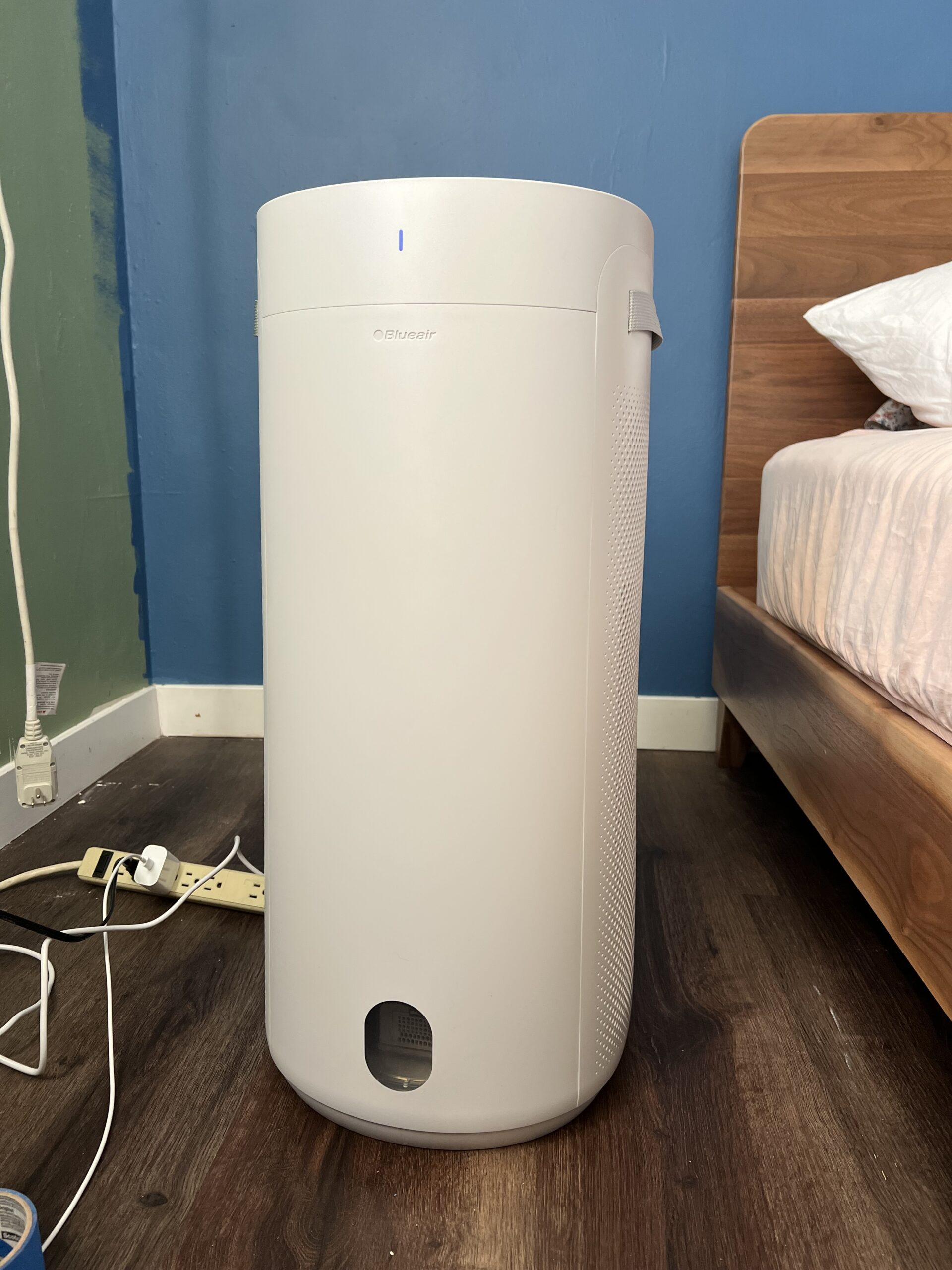 A white air purifier is placed on a wooden floor next to a bed, with power cords and a power strip nearby against a blue wall.