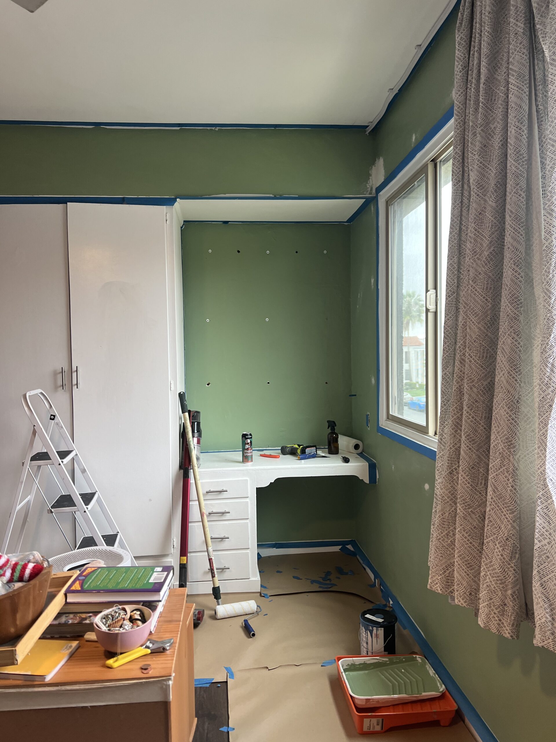 A small room in the process of being painted green, with painting supplies, a stepladder, covered furniture, and tape lining the edges of walls and ceiling.