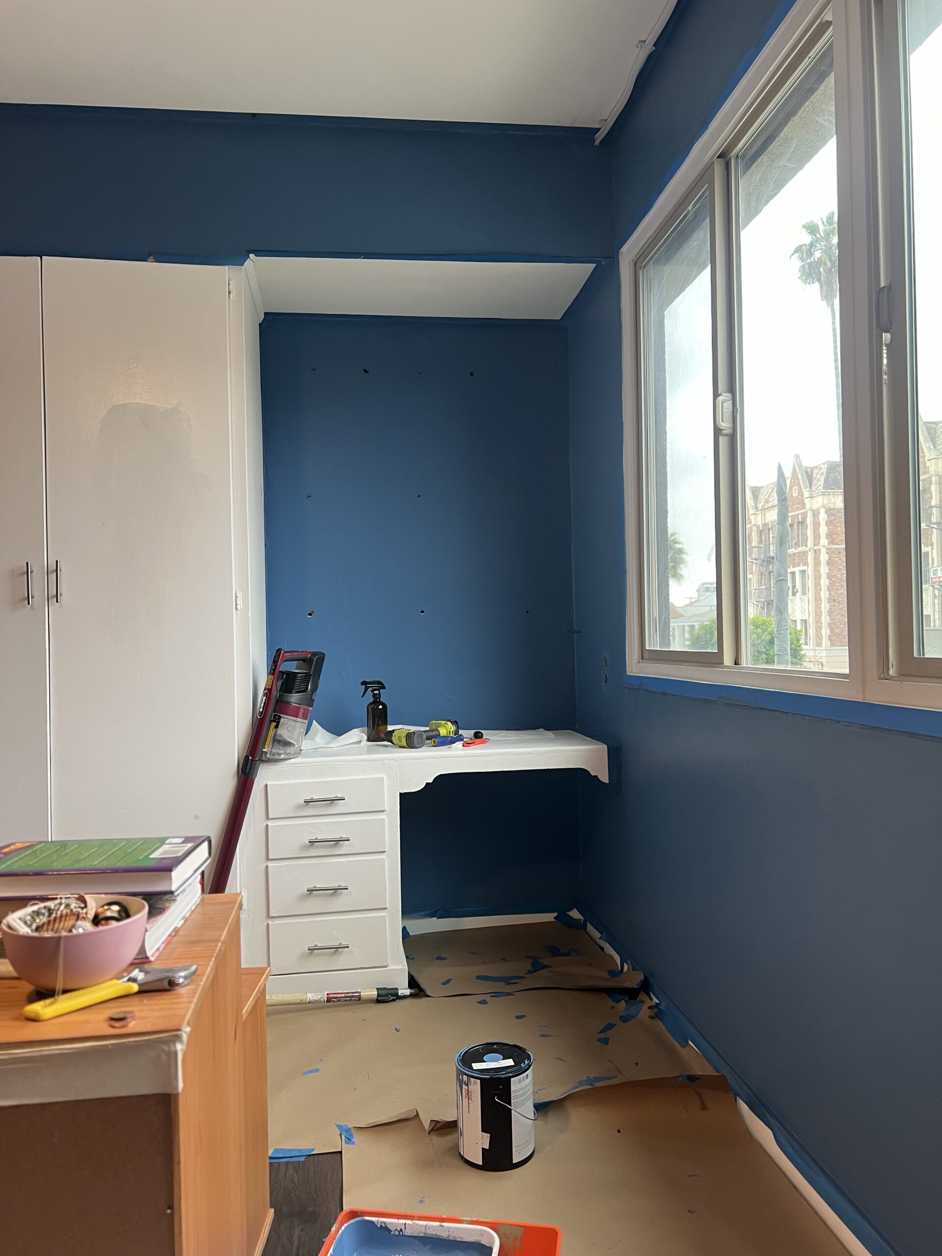 A small room being painted blue, with a white desk, paint can, tools, and painter’s tape on the floor. A window shows buildings and palm trees outside.