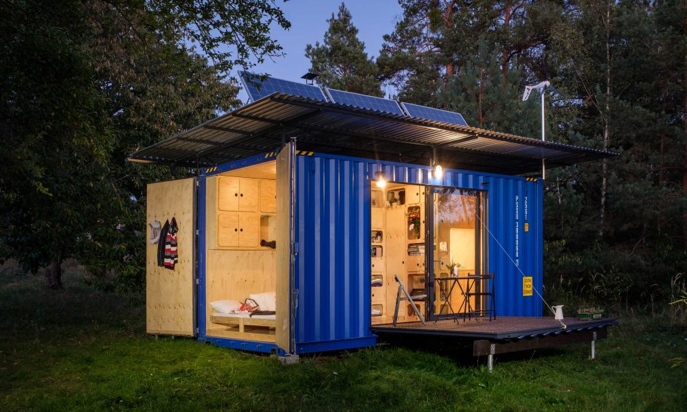 A blue shipping container converted into a tiny home with open doors, illuminated interior, wooden furnishings, work area, bed, and solar panels on the roof, set in a wooded area.