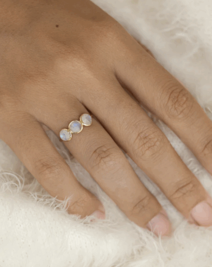 A hand wearing a gold ring set with three round, translucent stones, resting on a soft white textured surface.