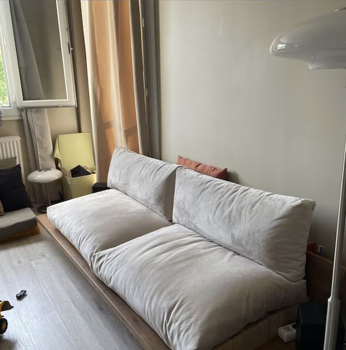 A beige cushioned sofa sits against a neutral wall in a room with wooden flooring, an open window, a lamp, and some scattered items on the floor.