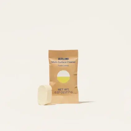 A brown paper packet labeled "Blueland Multi-Surface Cleaner Fresh Lemon" stands upright with a white cleaning tablet placed in front of it.