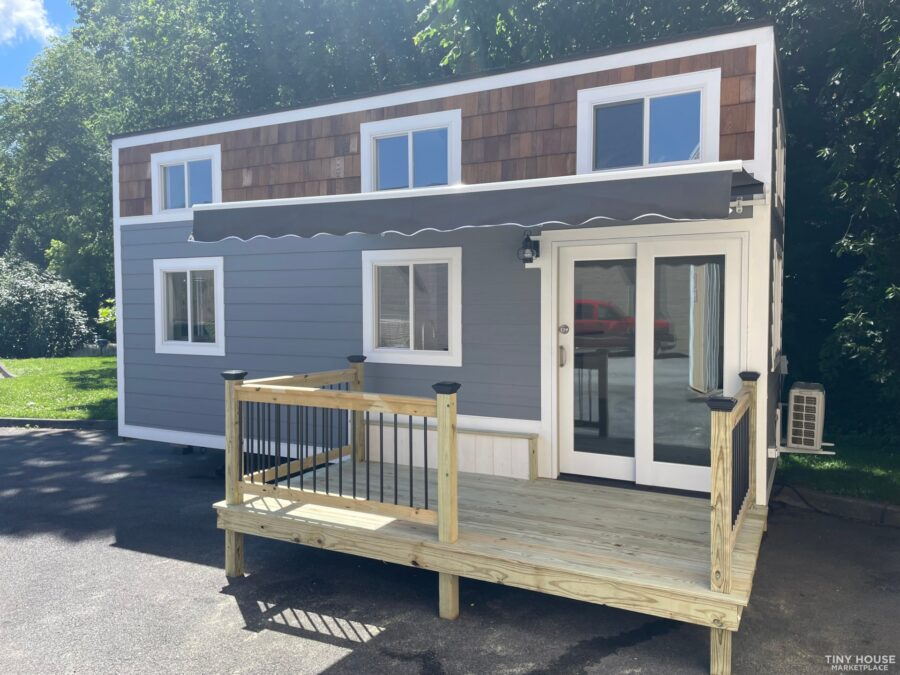 A small, modern house with a blue exterior, white trim, and a wooden porch with railing. It has an awning over the door and several windows, some with brown shingles above them.