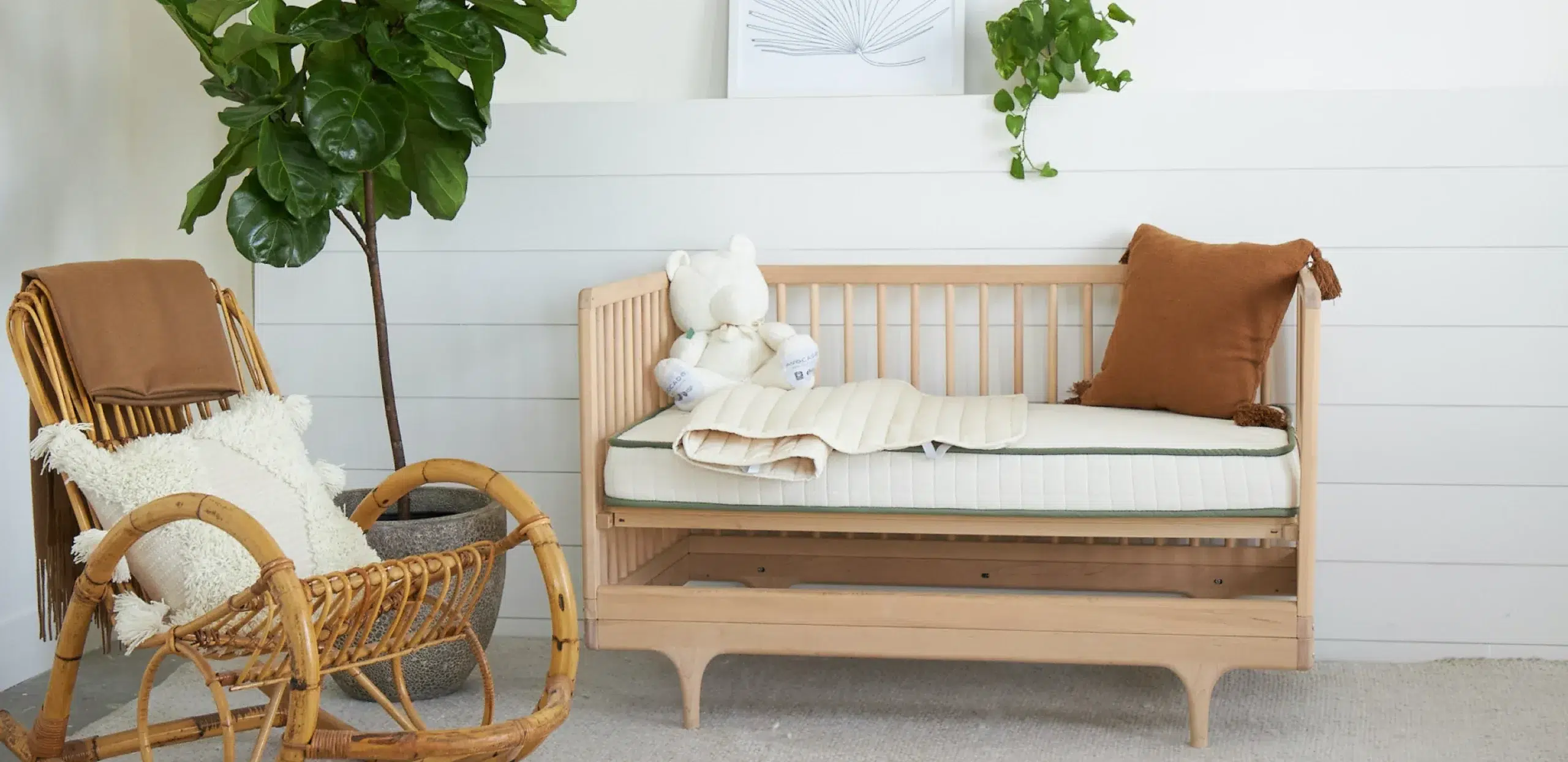 A wooden crib with a white mattress, brown pillow, and stuffed bear is beside a wicker chair and potted plants in a bright, minimal nursery.