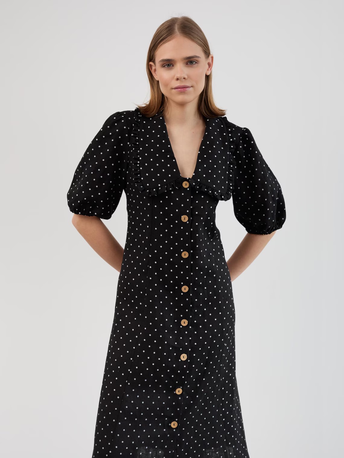 A woman wearing a black dress with white polka dots, short puff sleeves, and a row of buttons down the front, standing against a plain background.