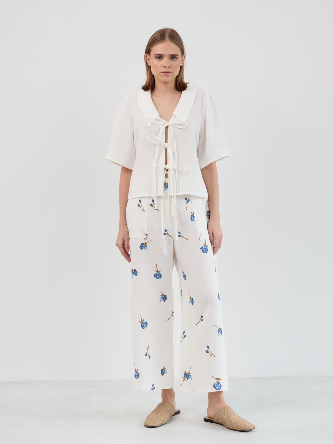 A person stands against a plain background wearing a white short-sleeve blouse with tie closures, white wide-leg pants with a blue floral print, and beige woven slip-on shoes.