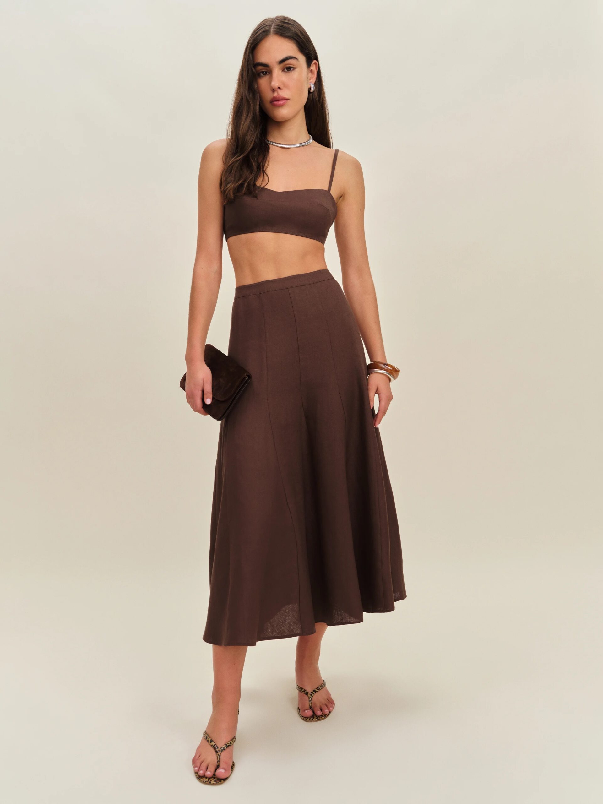 A woman stands wearing a brown crop top and matching midi skirt, holding a clutch bag, accessorized with sandals, bracelets, and a necklace against a plain background.