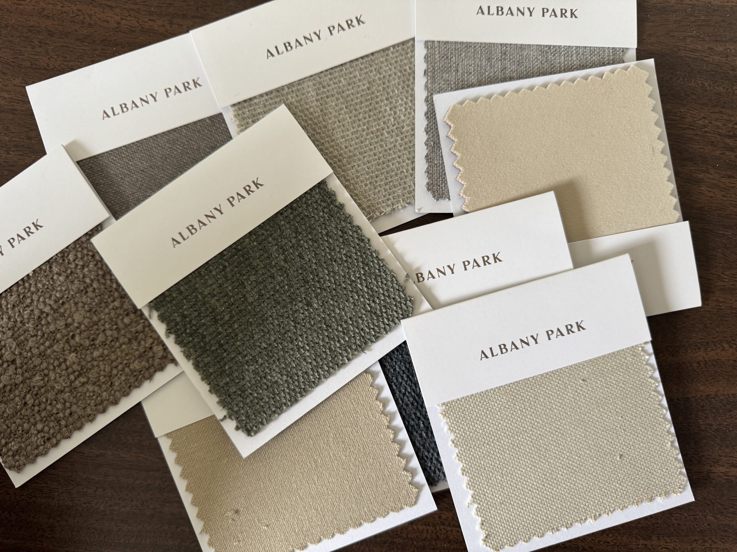 A scattered arrangement of Albany Park fabric swatches in various neutral tones, including beige, gray, and taupe, displayed on a dark wood surface.
