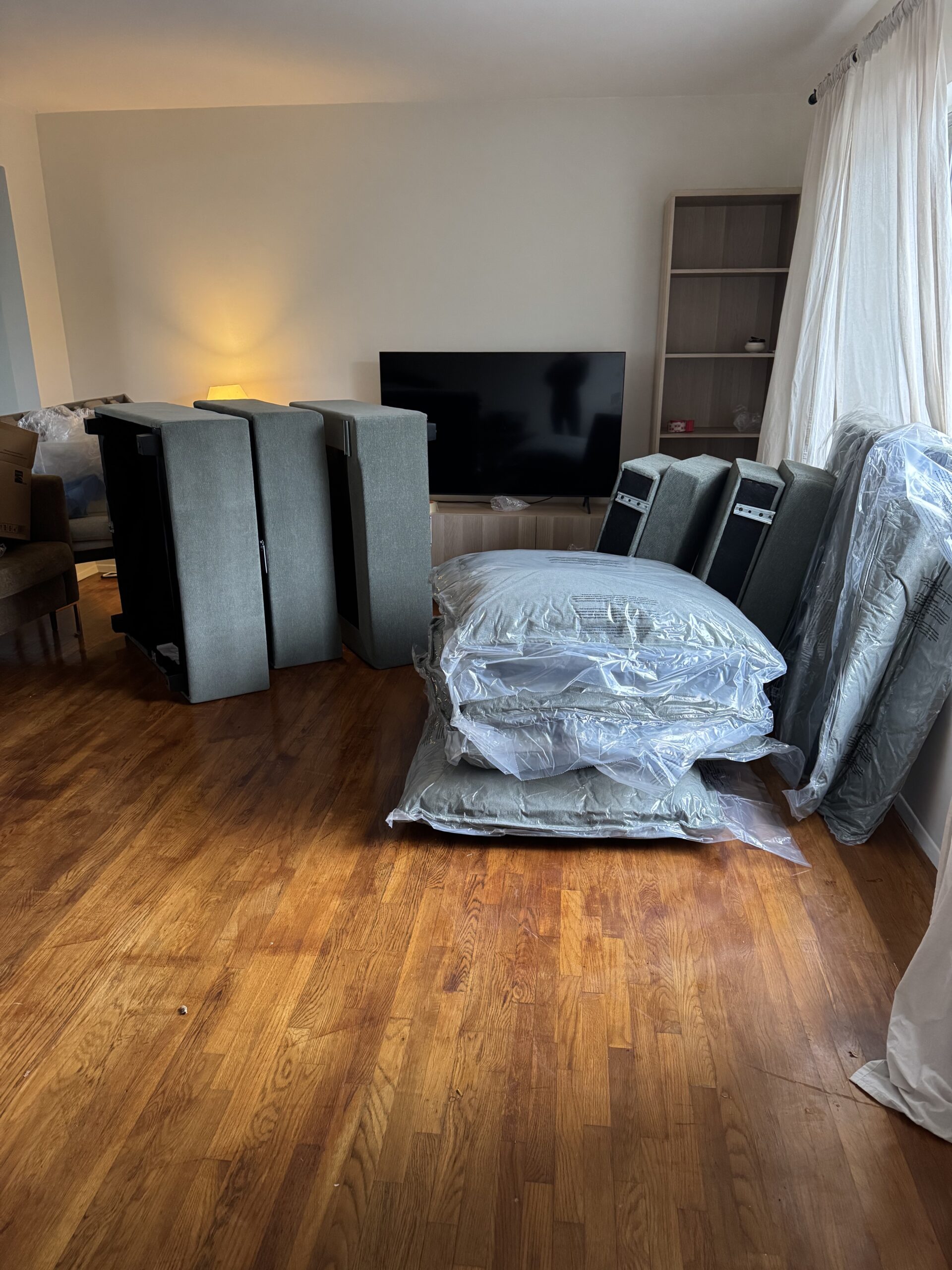 Couch cushions and parts wrapped in plastic are arranged on a wooden floor in a living room, with a TV and empty shelves in the background.