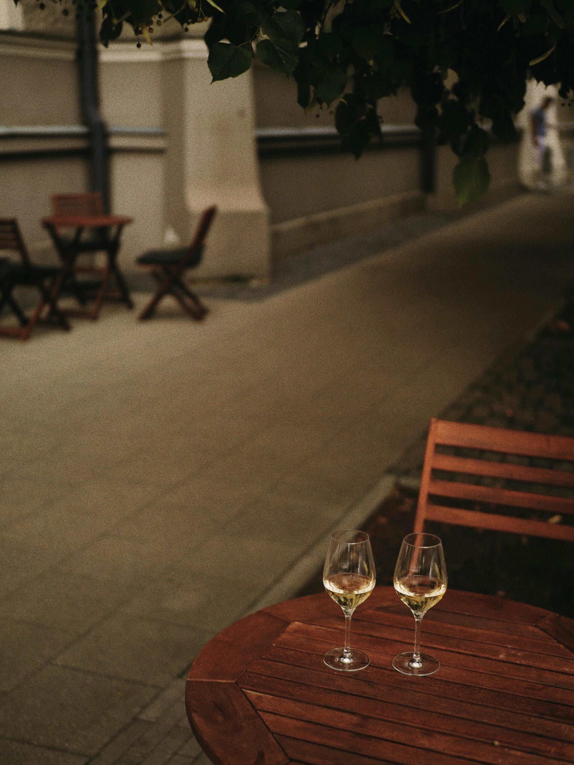 Two glasses of white wine on a wooden outdoor table, with empty chairs and tables along a quiet, shaded sidewalk.