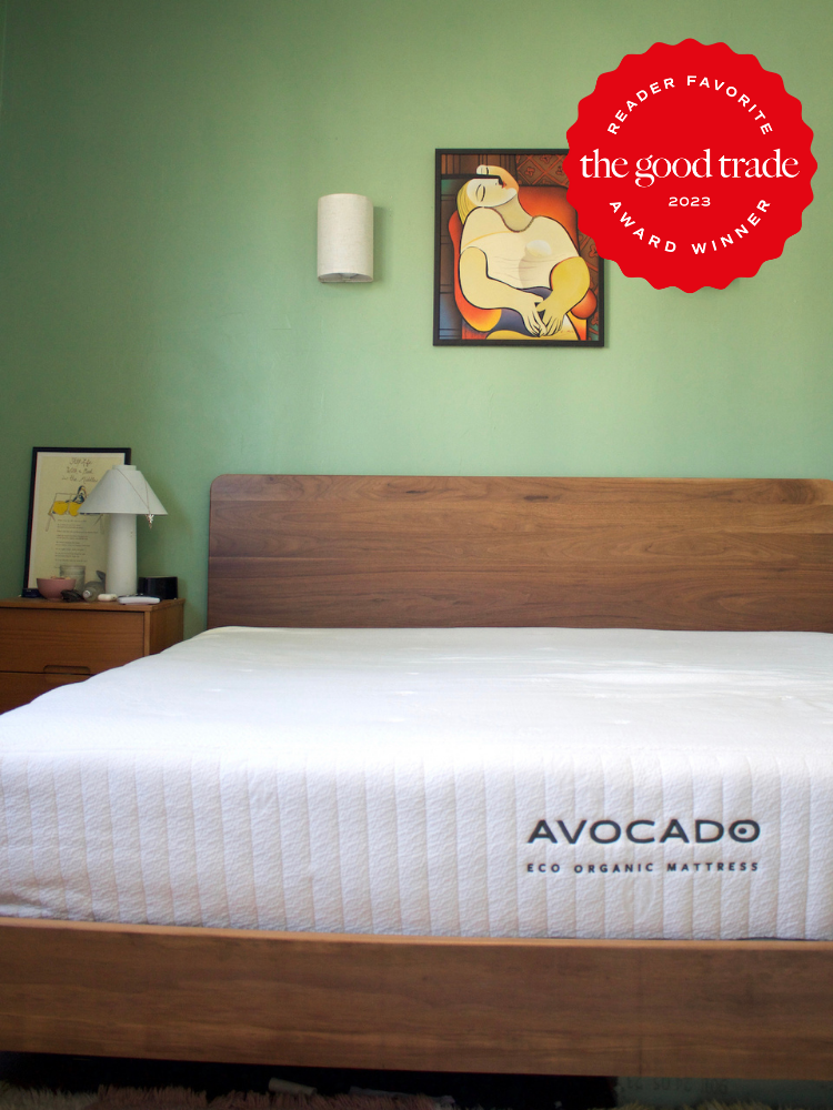 A bedroom with a wooden bed frame, an Avocado Eco Organic latex mattress, a nightstand with a lamp and picture, and a painting on a green wall. A red award seal is in the top right corner.