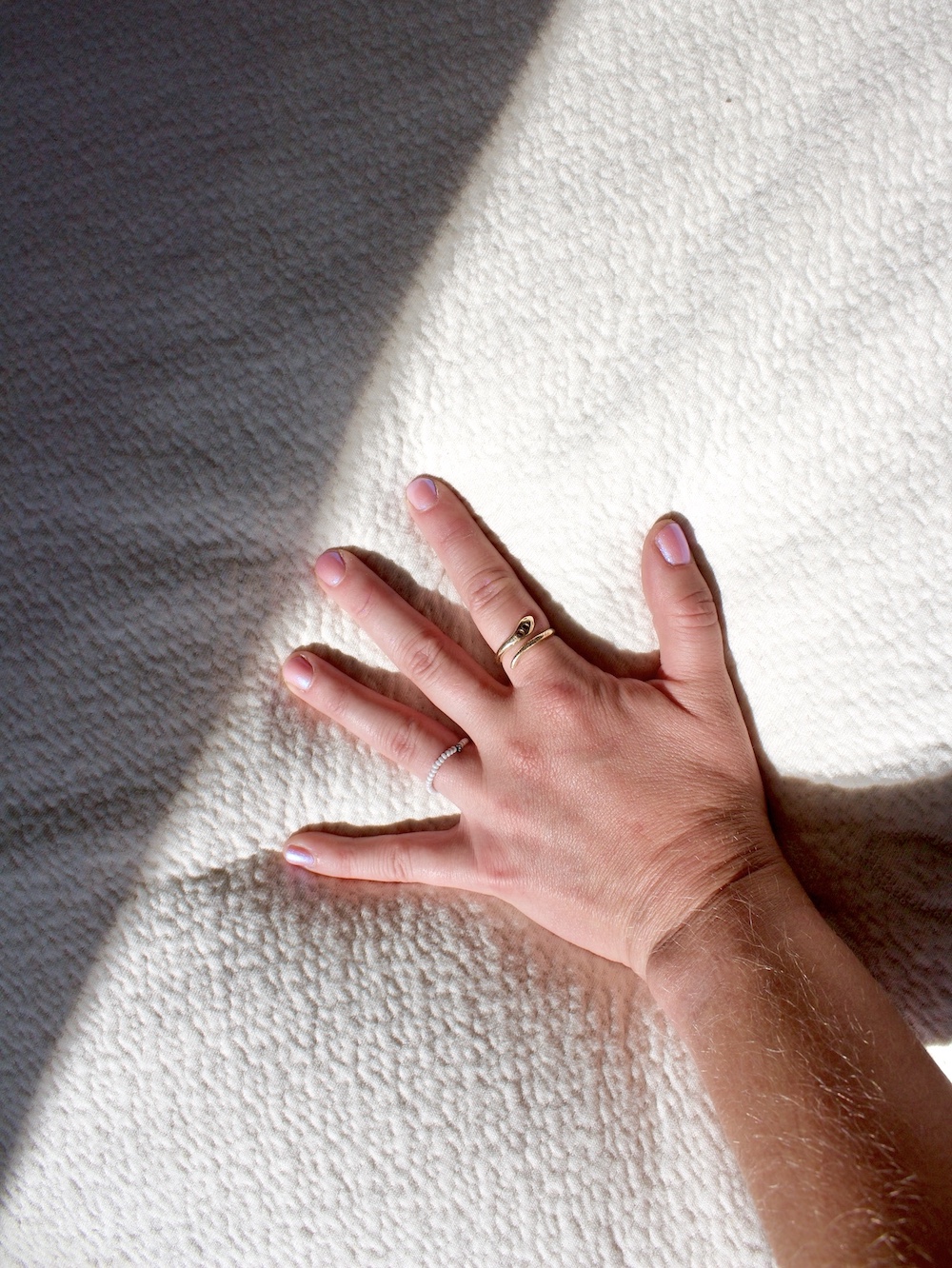 A hand with light pink nail polish and two rings rests on a textured white latex mattress in natural light.