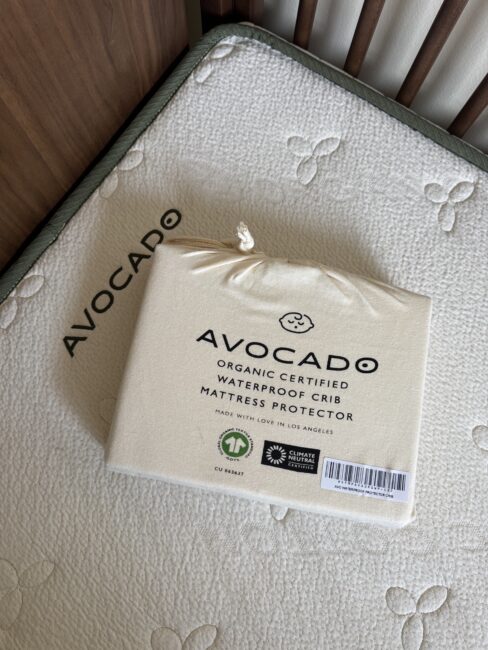 A packaged Avocado organic certified waterproof crib mattress protector rests on an Avocado crib mattress in a crib.