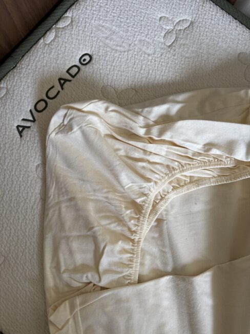 A fitted sheet with elastic corners is partially placed on an Avocado brand mattress with a textured surface and butterfly patterns.