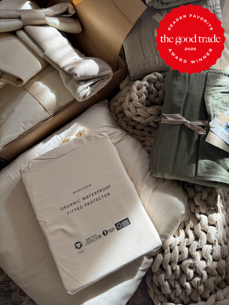 A variety of folded bedding items, including an Avocado organic waterproof fitted protector, displayed on a soft, textured surface with a red "The Good Trade Award Winner 2025" badge.