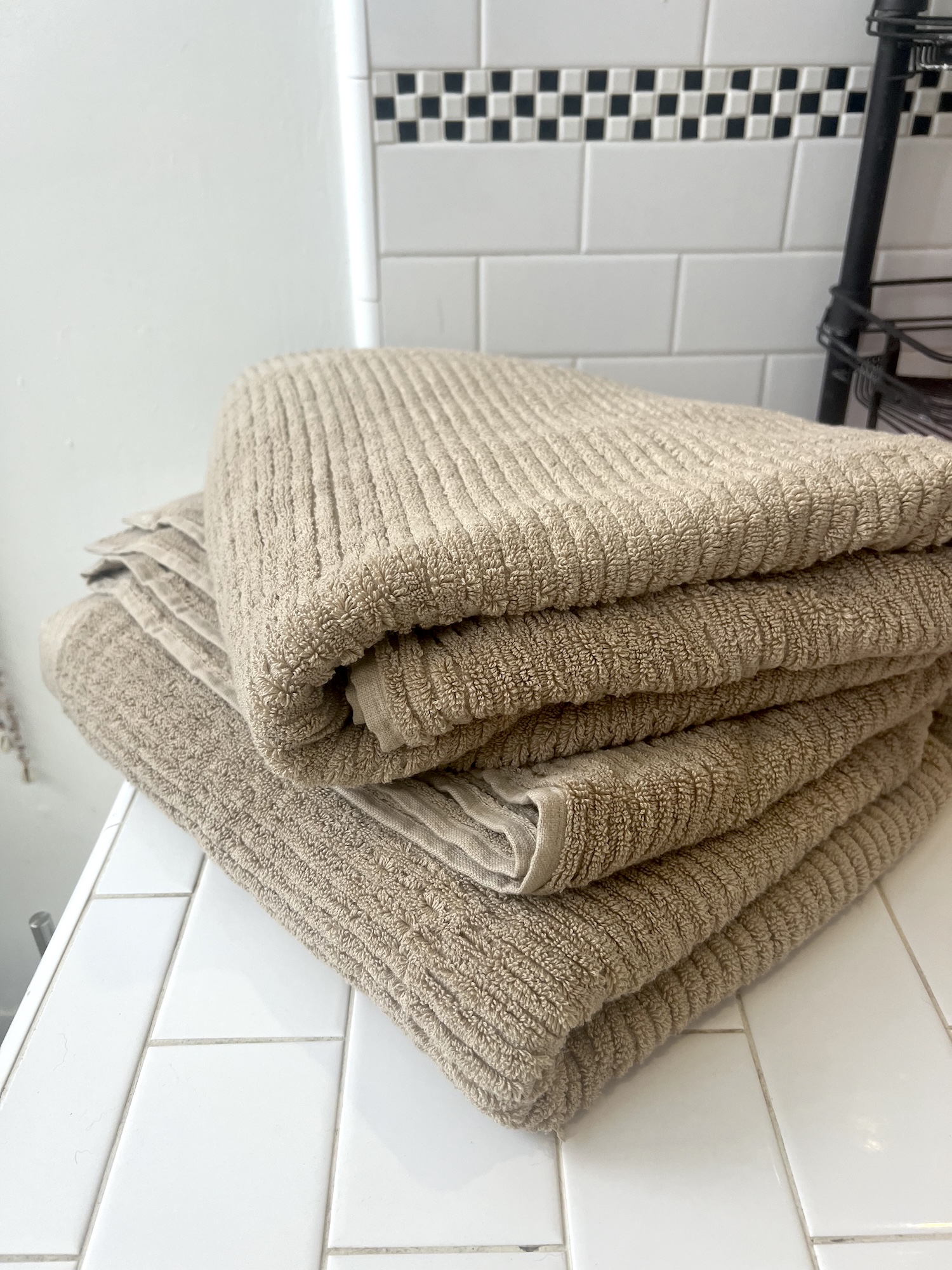 Three folded organic towels are stacked on a white tiled bathroom counter, with white and black tiled walls in the background.