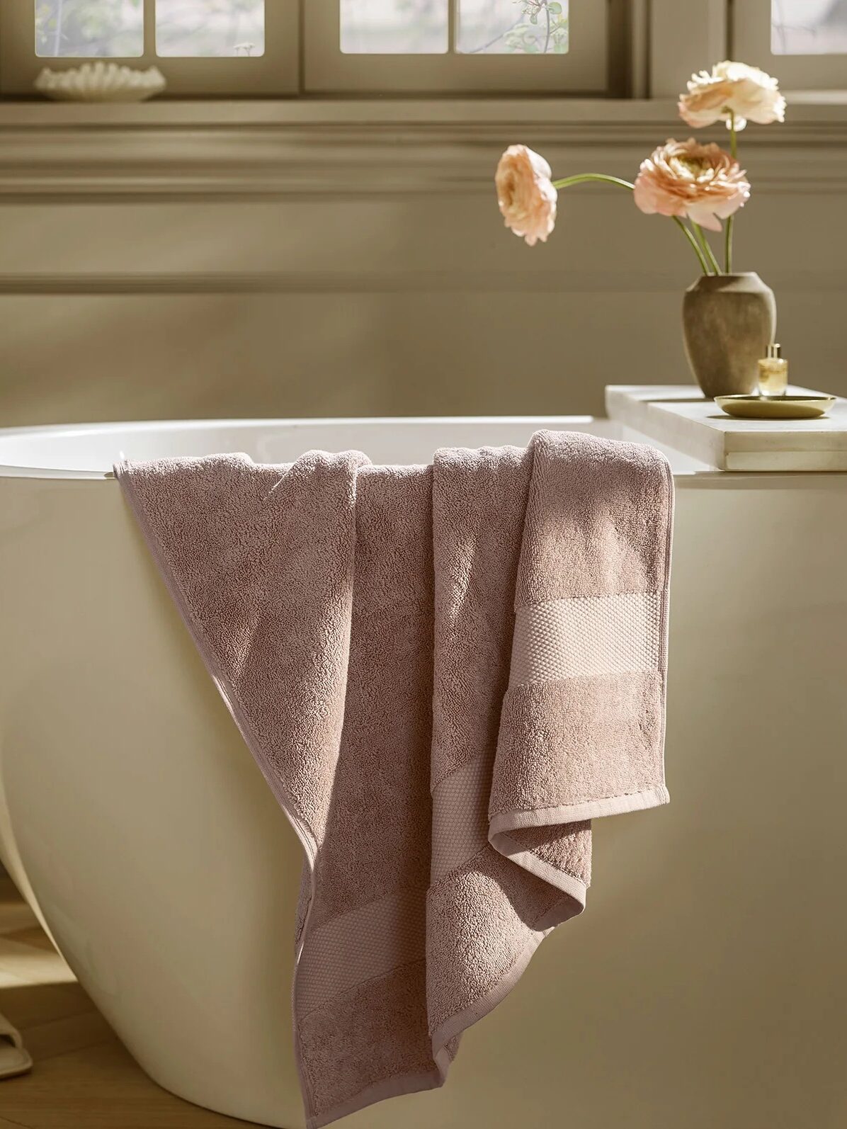 A folded mauve organic towel hangs over the edge of a white freestanding bathtub; a small vase with flowers and a candle sit on the tub’s ledge.