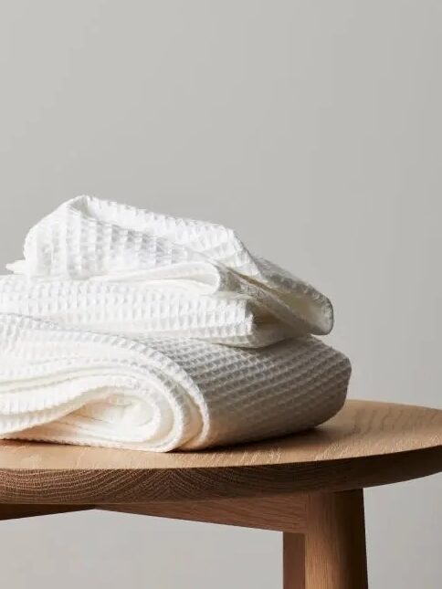 A folded white organic waffle weave towel rests on a wooden stool against a plain light gray background.