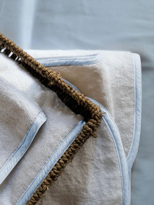 A close-up view of organic towels in beige with light blue edging and brown braided trim, neatly folded on a pale blue surface.