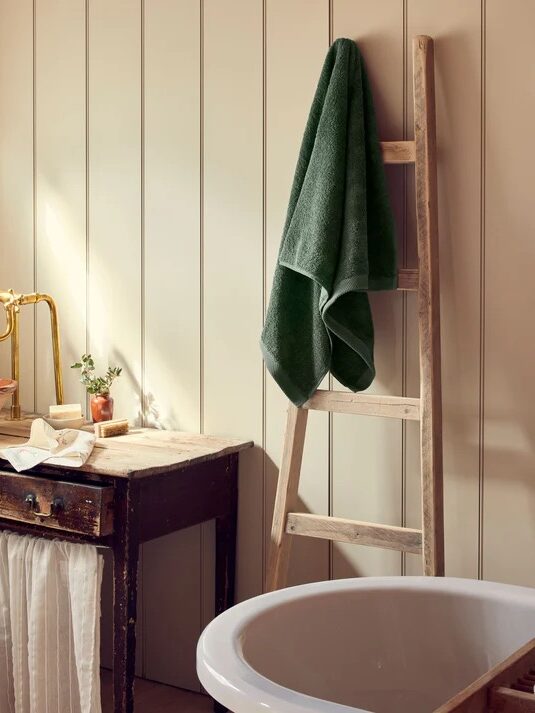 An organic towel hangs on a wooden ladder next to a round bathtub and a rustic wooden sink in a bathroom with vertical panel walls.
