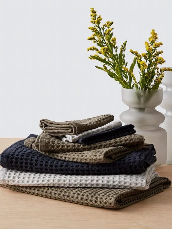 A stack of folded organic waffle-knit towels in white, green, and navy sits on a wooden surface next to a white vase with yellow flowers.