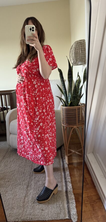 Person wearing a red floral dress and black shoes takes a mirror selfie in a living room with a plant, crib, and beige furniture visible in the background.