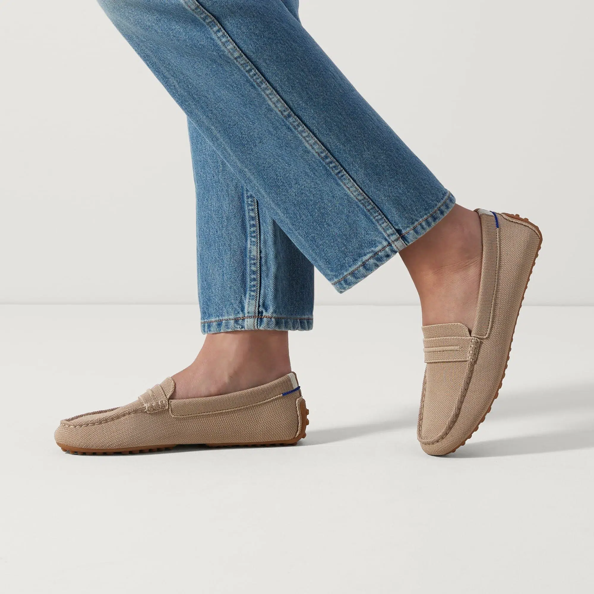 Person wearing light brown loafers with visible stitching and rubber soles, paired with blue jeans, walking on a plain white surface.