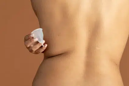 A person holds a white menstrual cup against their bare lower back, with a neutral brown background.
