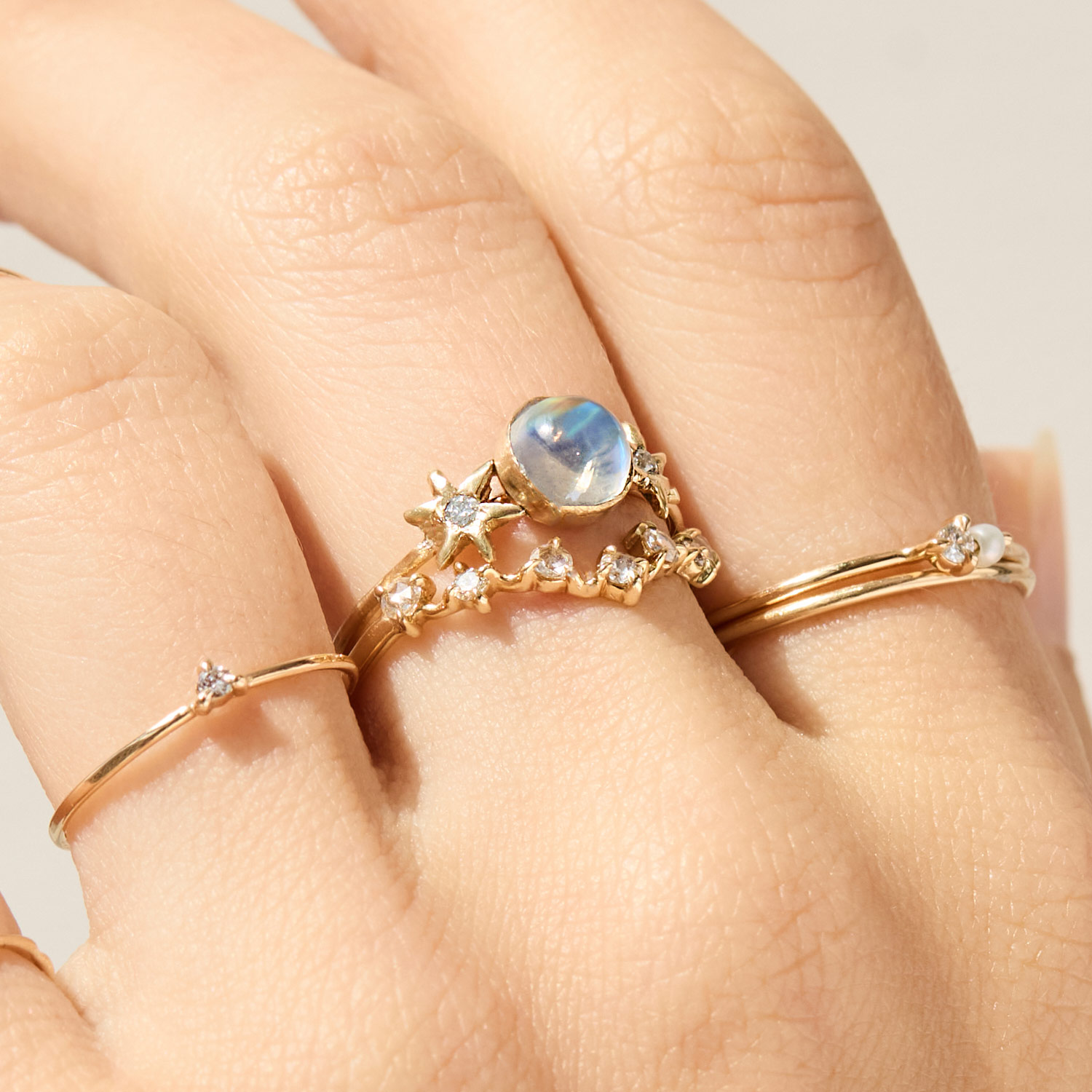 A hand wearing four gold rings, including a moonstone ring, a ring with star and diamond accents, a thin band with a small diamond, and a simple gold band.