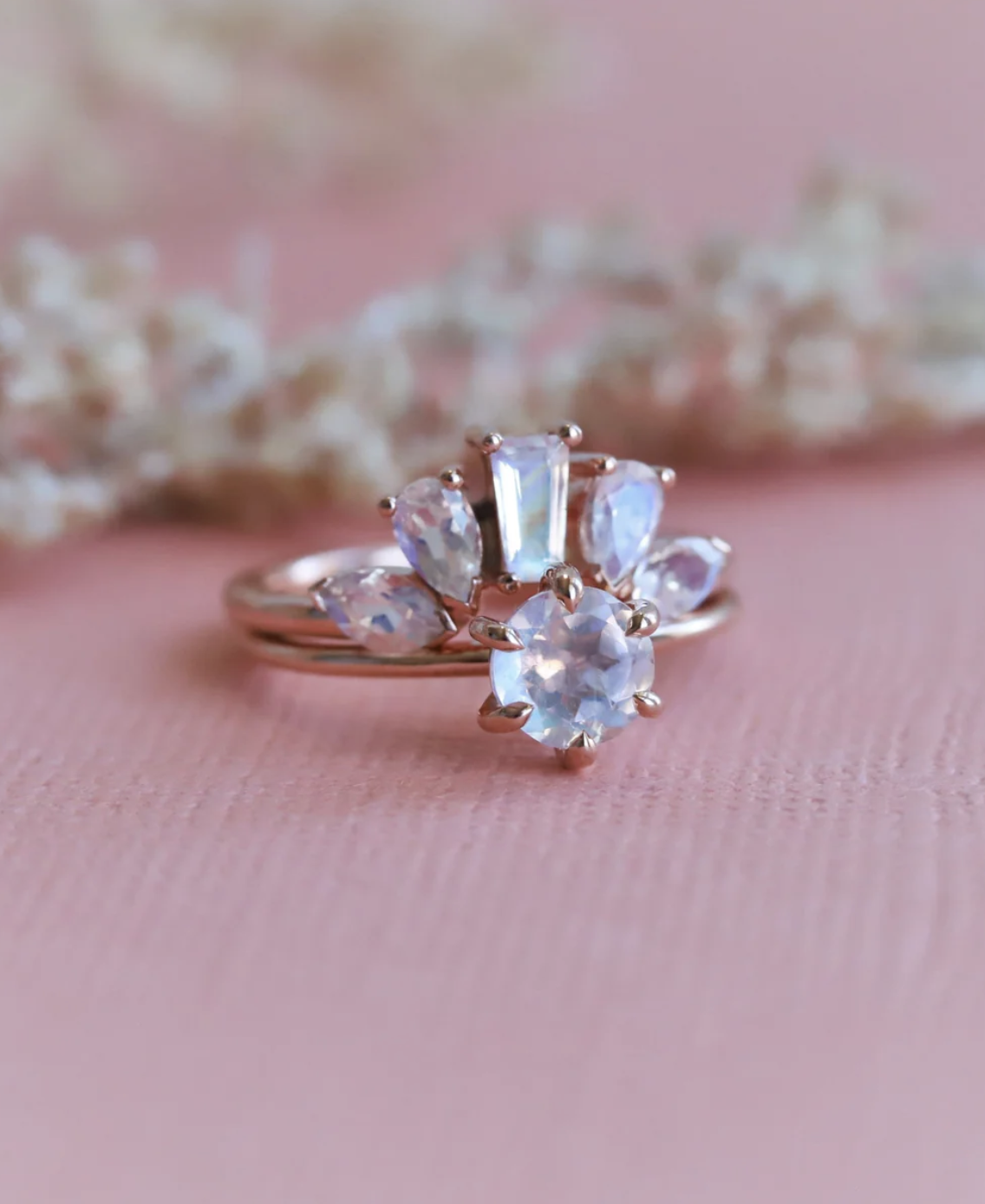 A rose gold ring set with a round central stone and five fan-shaped accent stones, displayed on a pink textured surface with blurred white flowers in the background.
