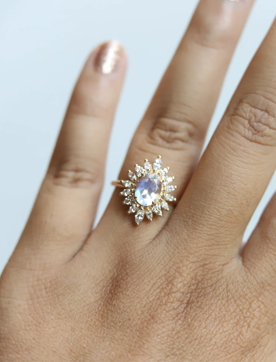 A hand with painted nails wears a gold ring featuring a large oval gemstone surrounded by small clear stones in a starburst pattern.