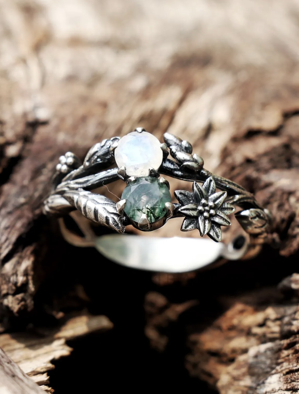 A silver ring with two round gemstones and floral, leaf, and vine details rests on textured wood.