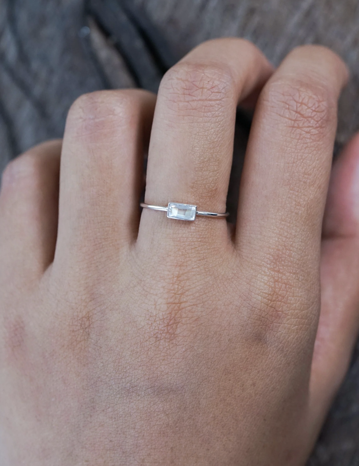 A hand wearing a thin silver ring with a small rectangular clear gemstone set horizontally on the ring finger.