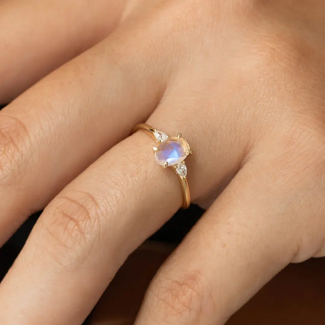 A hand wearing a gold ring with a central oval moonstone and two small clear side stones.