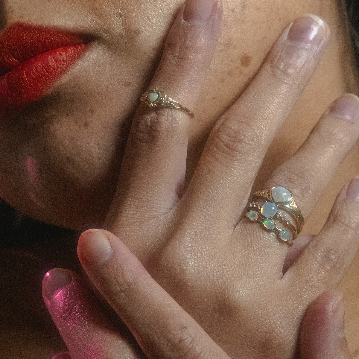 Close-up of a person with red lipstick wearing multiple gold rings with opal or moonstone gems on their fingers and hand touching their chin.